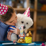 Young woman and her dog are wearing party hats and celebrating dogs 1st birthday at home.